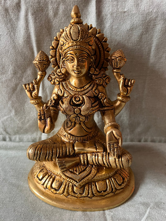 This brass Lakshmi statue from Saanskrityam Handicraft Store features the Goddess MahaLakshmi seated with four arms. She holds lotus flowers in two hands while displaying symbolic gestures with the others. Adorned with intricate jewelry and a serene expression, she embodies wealth and prosperity, making it an ideal choice for festive and auspicious gifts.
