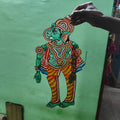 A hand displays a puppet from the Ramayana flat puppet set by Saanskrityam Handicraft Store, against a green wall. This elaborately decorated figure showcases vibrant colors—blue, red, and yellow—with intricate patterns and jewelry designs, potentially depicting the heroic Rama.