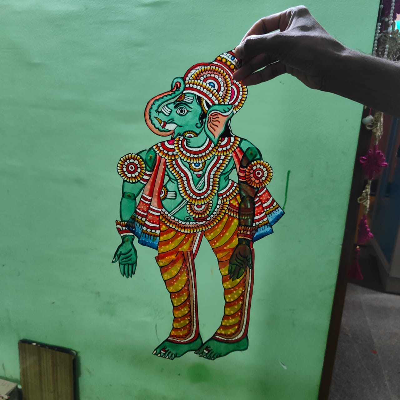 A hand displays a puppet from the Ramayana flat puppet set by Saanskrityam Handicraft Store, against a green wall. This elaborately decorated figure showcases vibrant colors—blue, red, and yellow—with intricate patterns and jewelry designs, potentially depicting the heroic Rama.