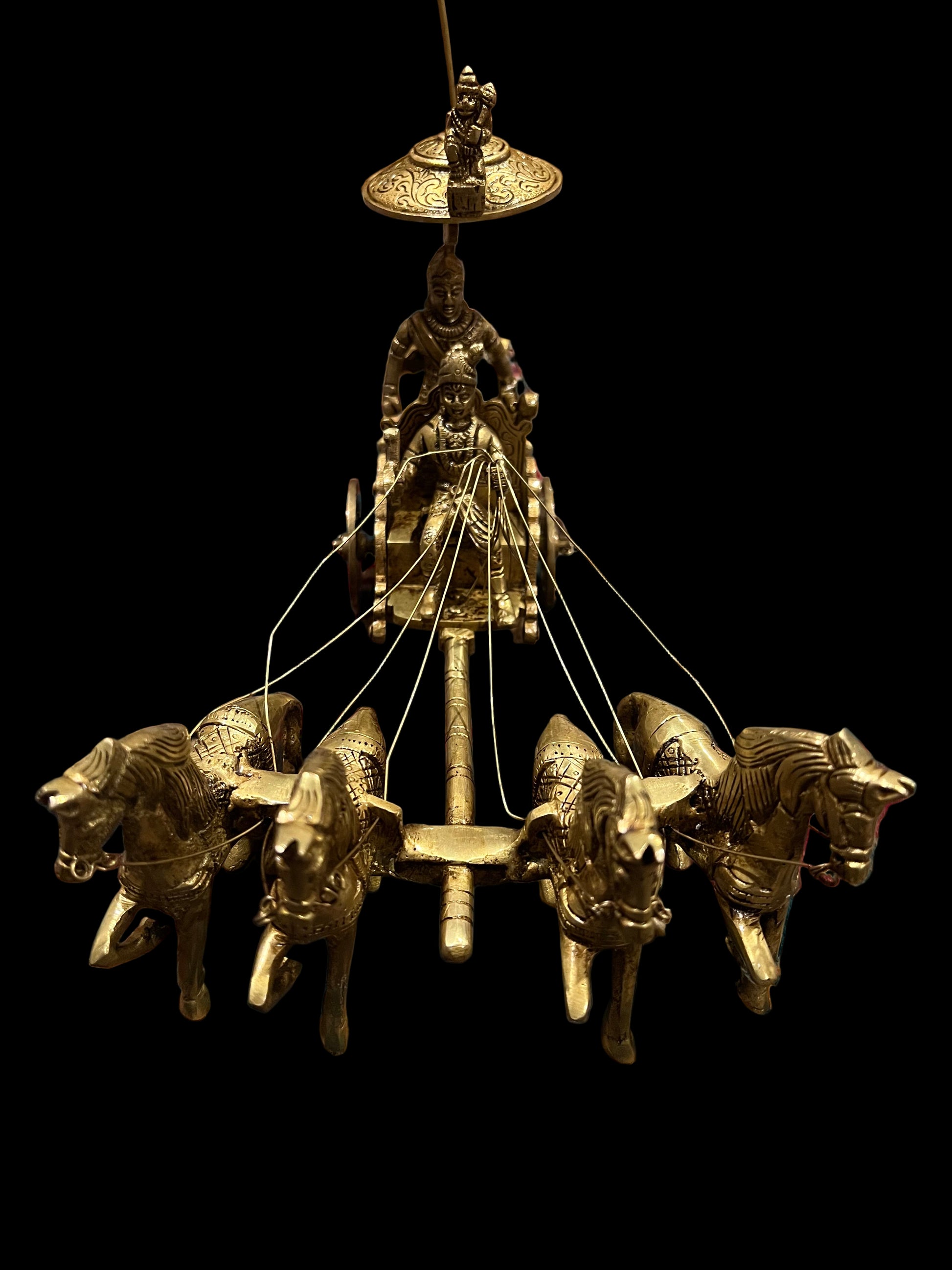 A bronze chariot sculpture named "Krishna and Arjuna / Bhagavad Gita at Kurukshetra," crafted by the Saanskrityam Handicraft Store, depicting four horses with Arjuna and Krishna holding a bow under an umbrella, is elegantly placed on an intricately patterned blue and red rug.