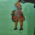 A hand displays a puppet from the Ramayana flat puppet set by Saanskrityam Handicraft Store, against a green wall. This elaborately decorated figure showcases vibrant colors—blue, red, and yellow—with intricate patterns and jewelry designs, potentially depicting the heroic Rama.