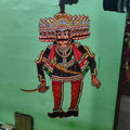 A hand displays a puppet from the Ramayana flat puppet set by Saanskrityam Handicraft Store, against a green wall. This elaborately decorated figure showcases vibrant colors—blue, red, and yellow—with intricate patterns and jewelry designs, potentially depicting the heroic Rama.