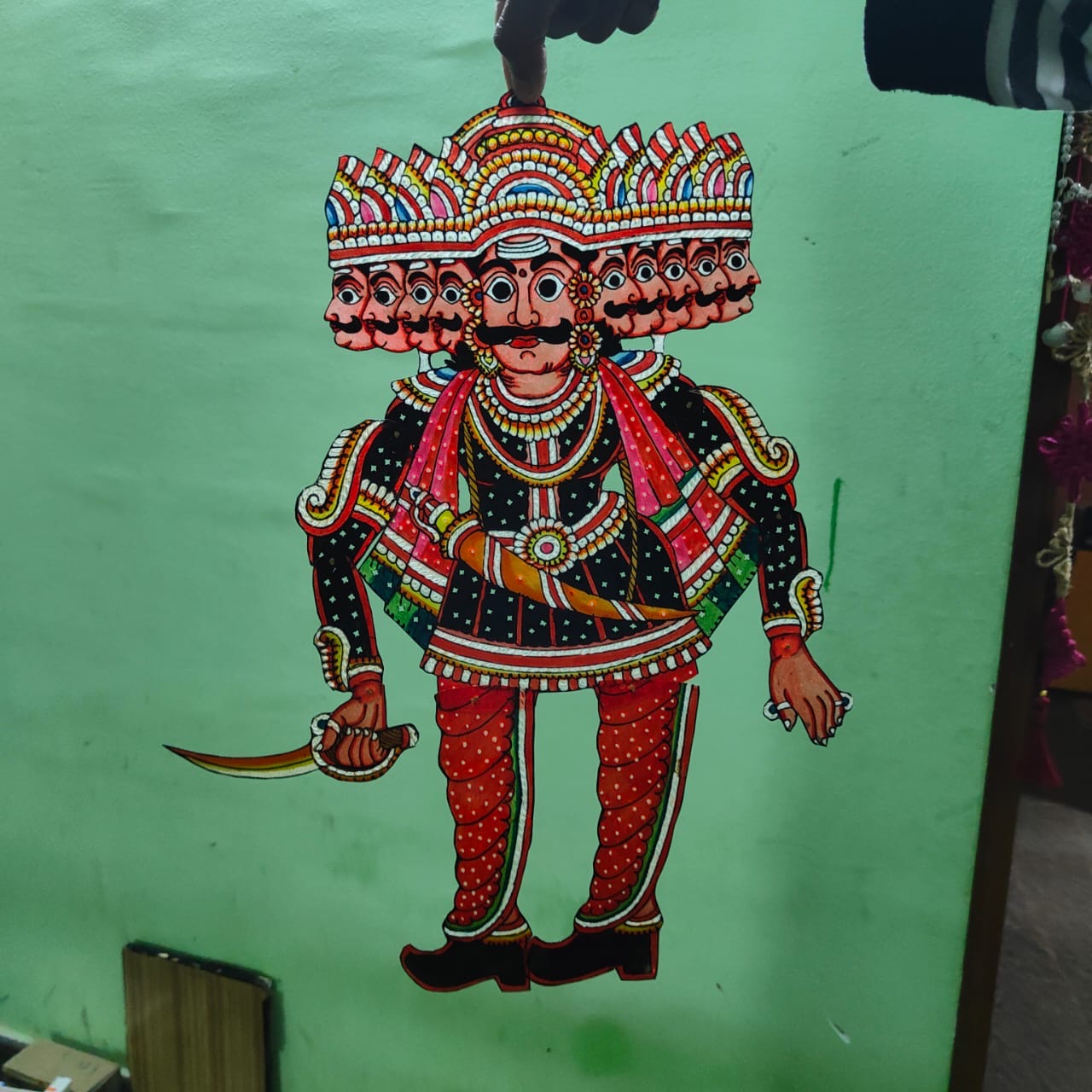 A hand displays a puppet from the Ramayana flat puppet set by Saanskrityam Handicraft Store, against a green wall. This elaborately decorated figure showcases vibrant colors—blue, red, and yellow—with intricate patterns and jewelry designs, potentially depicting the heroic Rama.