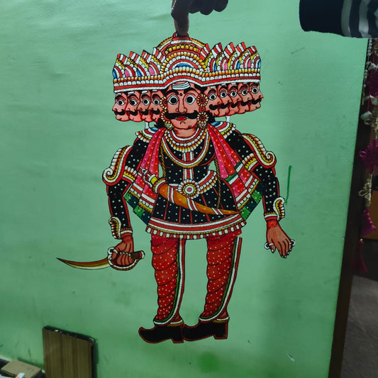 A hand displays a puppet from the Ramayana flat puppet set by Saanskrityam Handicraft Store, against a green wall. This elaborately decorated figure showcases vibrant colors—blue, red, and yellow—with intricate patterns and jewelry designs, potentially depicting the heroic Rama.