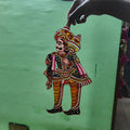 A hand displays a puppet from the Ramayana flat puppet set by Saanskrityam Handicraft Store, against a green wall. This elaborately decorated figure showcases vibrant colors—blue, red, and yellow—with intricate patterns and jewelry designs, potentially depicting the heroic Rama.