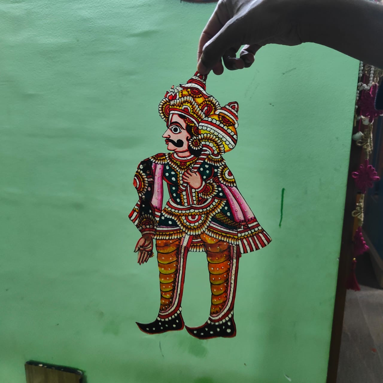 A hand displays a puppet from the Ramayana flat puppet set by Saanskrityam Handicraft Store, against a green wall. This elaborately decorated figure showcases vibrant colors—blue, red, and yellow—with intricate patterns and jewelry designs, potentially depicting the heroic Rama.