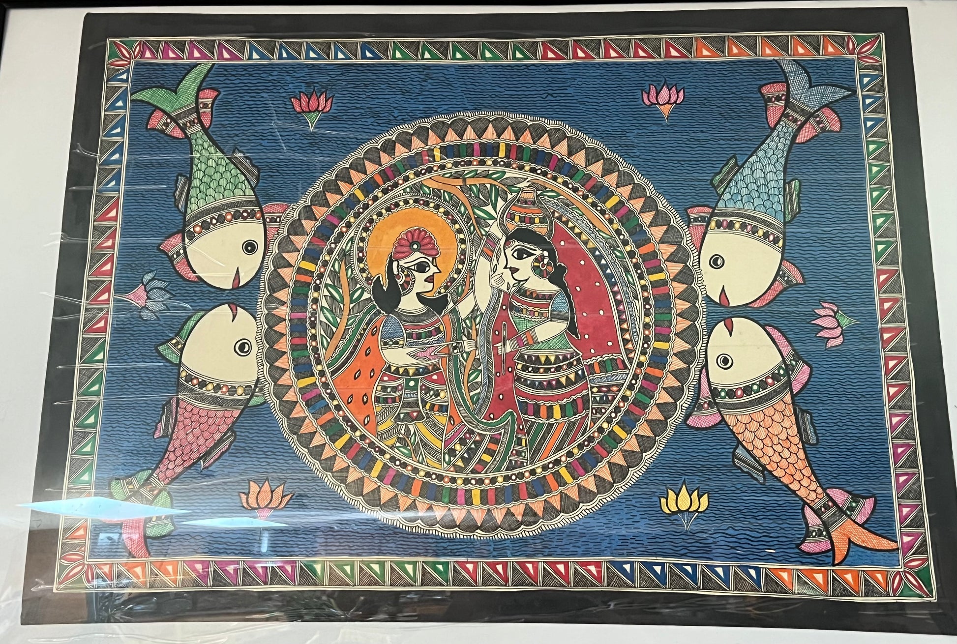 The Mithila or Madhubani style artwork painting from Saanskrityam Handicraft Store beautifully showcases two figures in lively traditional attire, facing each other. One figure is depicted with a yellow face and the other with a blue one, both embellished with intricate patterns and floral motifs. They stand beneath a tree featuring red and green leaves, encapsulating the essence of this folk art style.