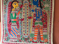 The Mithila or Madhubani style artwork painting from Saanskrityam Handicraft Store beautifully showcases two figures in lively traditional attire, facing each other. One figure is depicted with a yellow face and the other with a blue one, both embellished with intricate patterns and floral motifs. They stand beneath a tree featuring red and green leaves, encapsulating the essence of this folk art style.