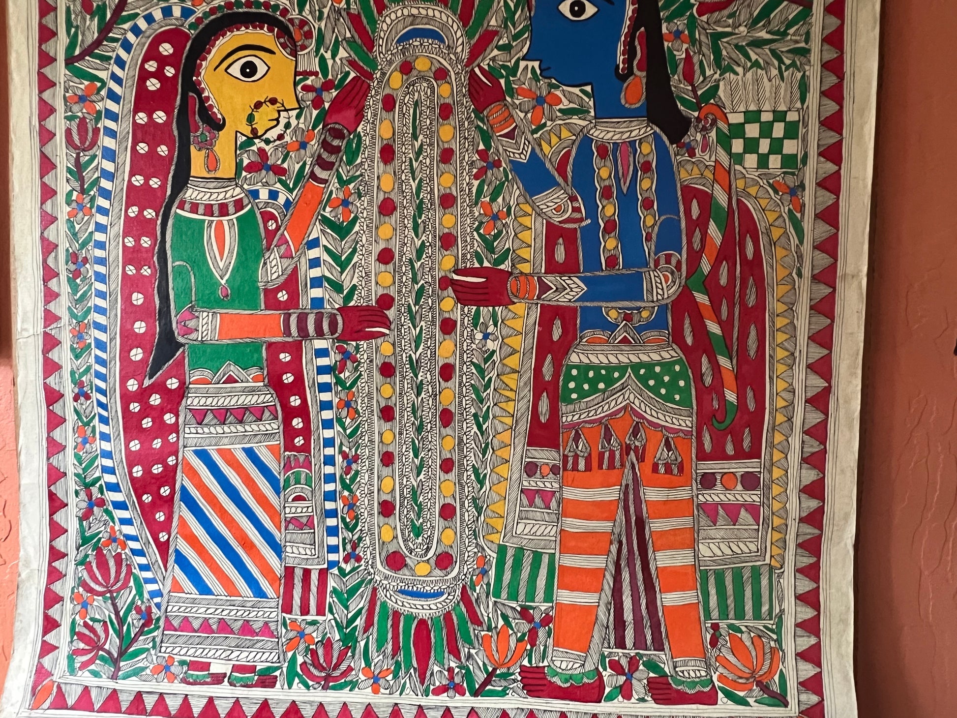 The Mithila or Madhubani style artwork painting from Saanskrityam Handicraft Store beautifully showcases two figures in lively traditional attire, facing each other. One figure is depicted with a yellow face and the other with a blue one, both embellished with intricate patterns and floral motifs. They stand beneath a tree featuring red and green leaves, encapsulating the essence of this folk art style.