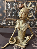 A bronze Chhattisgarh art sculpture from the Saanskrityam Handicraft Store presents a seated tribal woman, adorned with intricate jewelry and a headdress, holding a small object. The background includes a decorative metal panel embellished with geometric patterns and a sun motif.