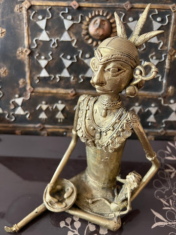 A bronze Chhattisgarh art sculpture from the Saanskrityam Handicraft Store presents a seated tribal woman, adorned with intricate jewelry and a headdress, holding a small object. The background includes a decorative metal panel embellished with geometric patterns and a sun motif.