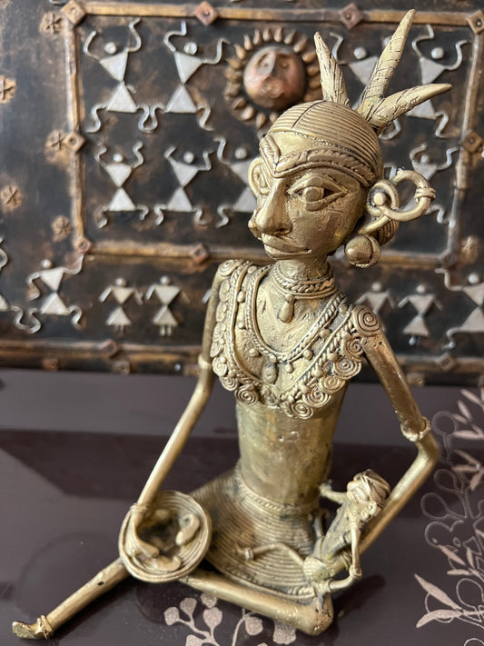 A bronze Chhattisgarh art sculpture from the Saanskrityam Handicraft Store presents a seated tribal woman, adorned with intricate jewelry and a headdress, holding a small object. The background includes a decorative metal panel embellished with geometric patterns and a sun motif.