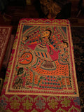 The Mithila or Madhubani style artwork painting from Saanskrityam Handicraft Store beautifully showcases two figures in lively traditional attire, facing each other. One figure is depicted with a yellow face and the other with a blue one, both embellished with intricate patterns and floral motifs. They stand beneath a tree featuring red and green leaves, encapsulating the essence of this folk art style.