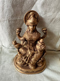 This brass Lakshmi statue from Saanskrityam Handicraft Store features the Goddess MahaLakshmi seated with four arms. She holds lotus flowers in two hands while displaying symbolic gestures with the others. Adorned with intricate jewelry and a serene expression, she embodies wealth and prosperity, making it an ideal choice for festive and auspicious gifts.