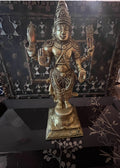 The Kartikeya statue from the Saanskrityam Handicraft Store's Tamilnadu brassware and bronzes collection features a serene depiction of the Hindu God. The brass figure is positioned on a pedestal, holding symbolic items in each hand, while his divine companion—a peacock—stands elegantly beside him, all set against a plain beige background.