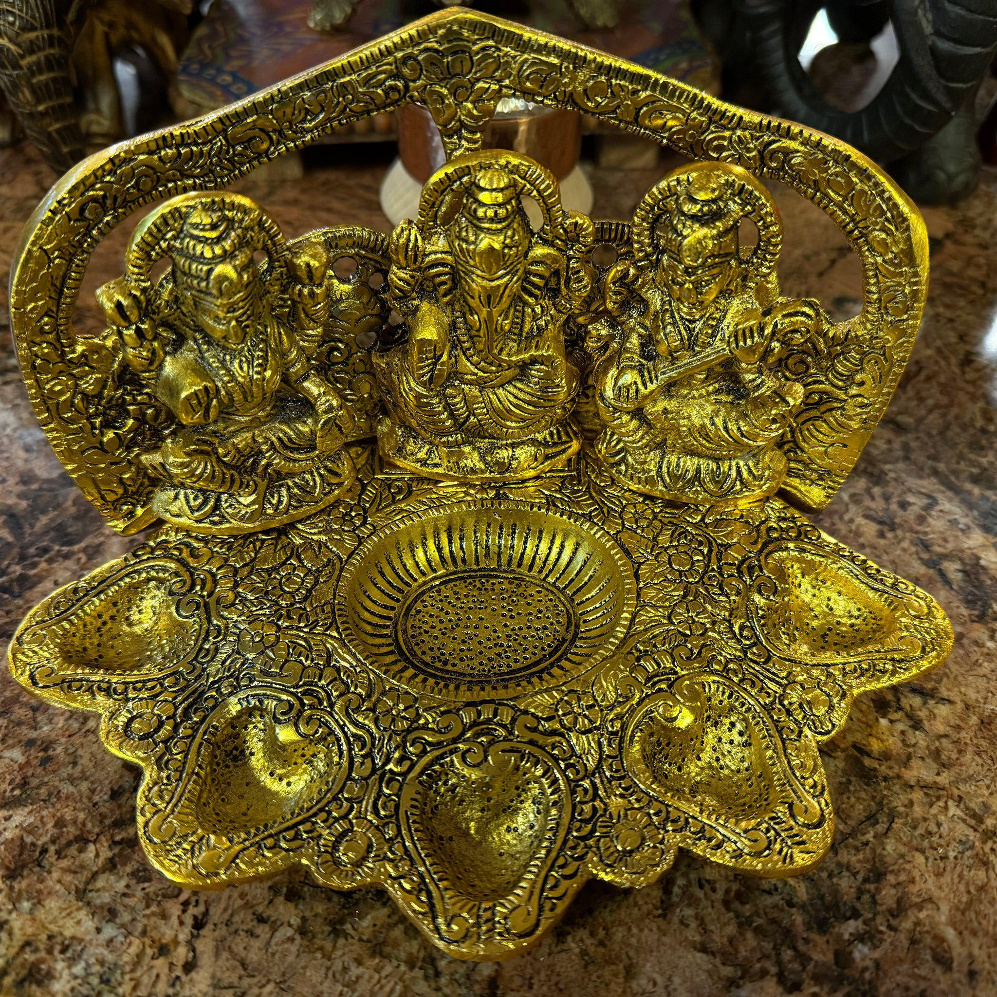 Ornate golden diya holder featuring three intricately carved deities seated on a textured surface, with leaf-shaped oil lamp holders surrounding a central bowl. Set on a brown stone table.