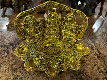 Ornate golden diya holder featuring three intricately carved deities seated on a textured surface, with leaf-shaped oil lamp holders surrounding a central bowl. Set on a brown stone table.