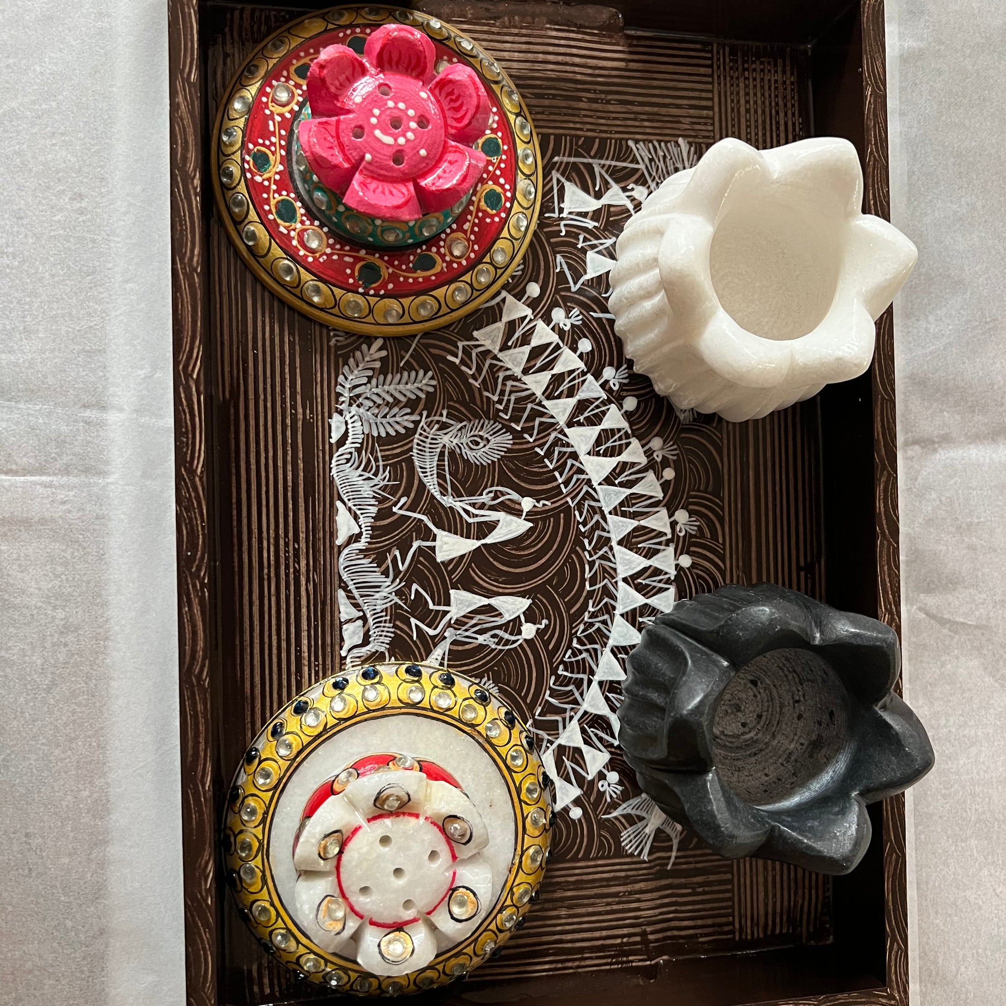 A decorative tray with two ornate round containers and two empty flower-shaped candle holders, one white and one black, placed on a white surface. The tray features intricate patterns inside.