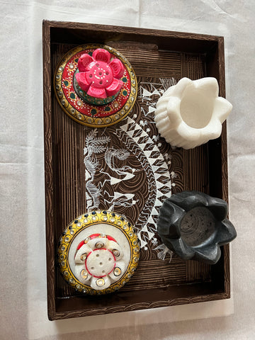 A decorative tray with two ornate round containers and two empty flower-shaped candle holders, one white and one black, placed on a white surface. The tray features intricate patterns inside.