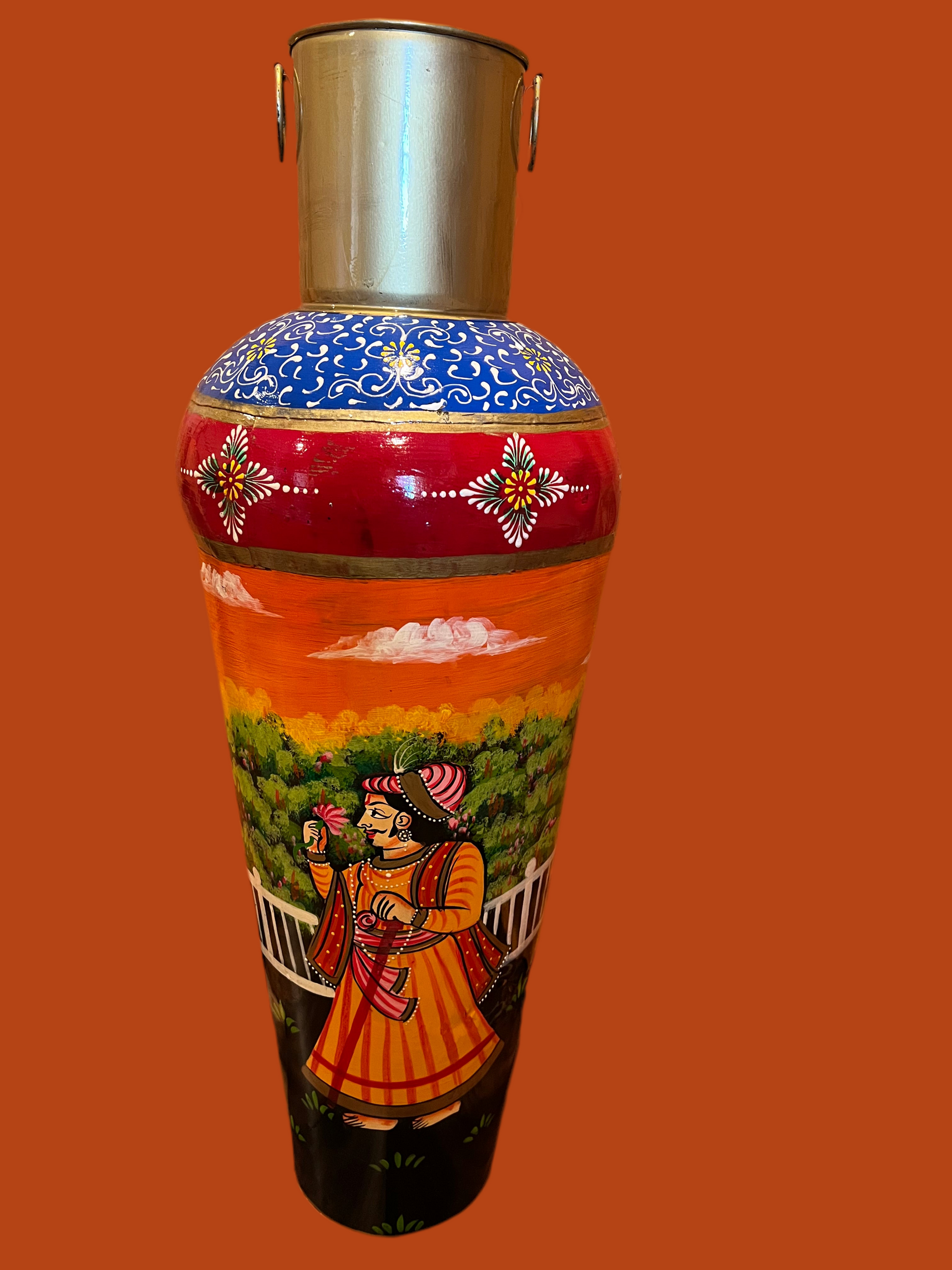 Experience the vibrant beauty of Saanskrityam Handicraft Store's "Rajasthani Art on Metal Vase." This handcrafted piece from India features colorful traditional artwork, with a central image depicting a man and woman in traditional attire riding a camel and intricate patterns in various bright colors surrounding them.