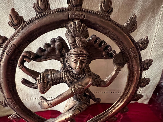 Shiva statue from the Tamil Nadu brassware Shivoham collection, part of Saanskrityam Handicraft Store, featuring the Hindu deity in a cosmic dance pose encircled by a ring of flames. This intricately detailed brass figure rests on a red and gold patterned cloth background.