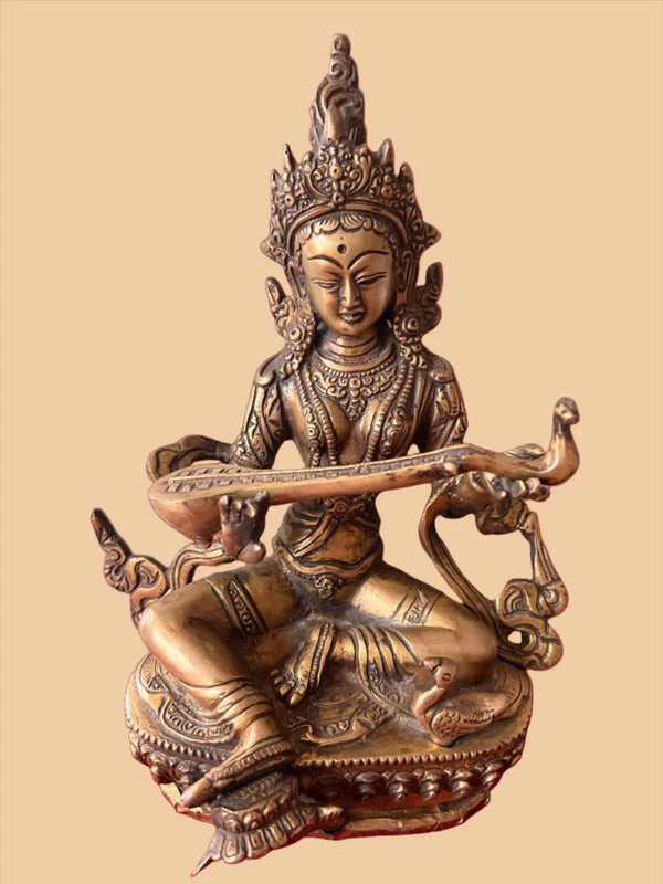 The 10-inch Saraswathi bronze statue from Saanskrityam Handicraft Store features a seated deity adorned with an ornate headdress and jewelry, holding a long, curved object, set against a plain background.