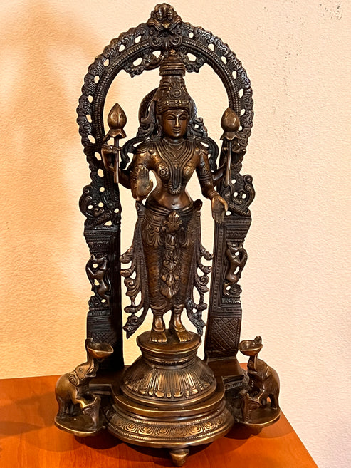 A 12-inch Goddess Lakshmi statue crafted by Saanskrityam Handicraft Store, showcasing the deity in traditional attire with an intricate headpiece and ornaments, framed by an ornate arch. The base features two elephants and rests on a wooden surface.