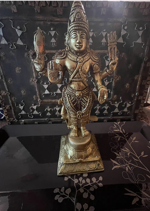 Introducing the 12-inch Kartikeya bronze statue by Saanskrityam Handicraft Store: a majestic figure intricately detailed and adorned with jewelry and a headdress, holding symbolic objects. This statue is set on a dark surface featuring a vine-like pattern, with a backdrop of a textured metallic screen.