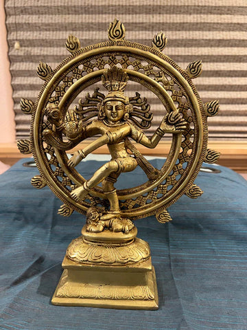 The 12-inch Nataraja bronze from Saanskrityam Handicraft Store is elegantly positioned on a square base, featuring a dancing figure encircled by flames, representing the cosmic dance. The statue boasts multiple arms, intricate details, and a serene expression, all beautifully set against a blue cloth background.