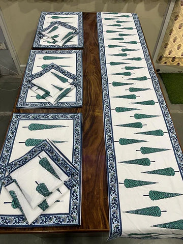 A wooden table is adorned with a matching fabric table runner, placemats, and napkins from Saanskrityam Handicraft Store. The fabric showcases green tree designs with blue floral borders, creating a cohesive and decorative dining setup.