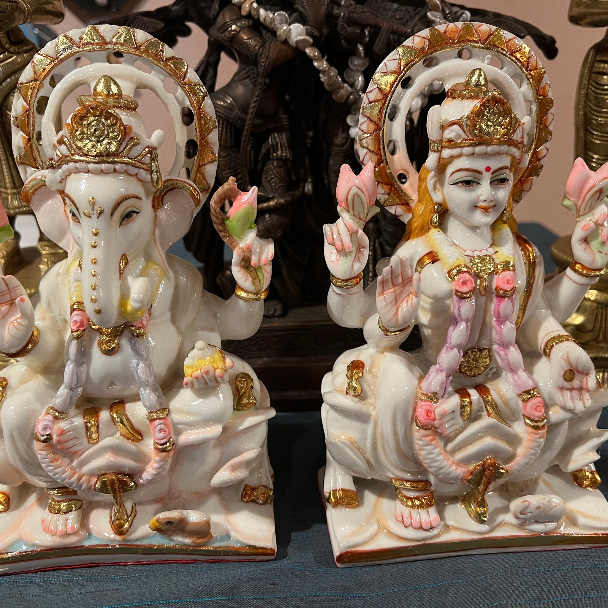 The Lakshmi Ganesh statues by Saanskrityam Handicraft Store feature two ornate marble figures representing Hindu deities. Ganesh is masterfully crafted with an elephant head, a crown, and holds symbolic items such as a lotus. Lakshmi is depicted seated, with multiple arms holding lotuses. Both statues are beautifully adorned in colorful and intricate attire with gold accents.