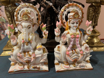 The Lakshmi Ganesh statues by Saanskrityam Handicraft Store feature two ornate marble figures representing Hindu deities. Ganesh is masterfully crafted with an elephant head, a crown, and holds symbolic items such as a lotus. Lakshmi is depicted seated, with multiple arms holding lotuses. Both statues are beautifully adorned in colorful and intricate attire with gold accents.