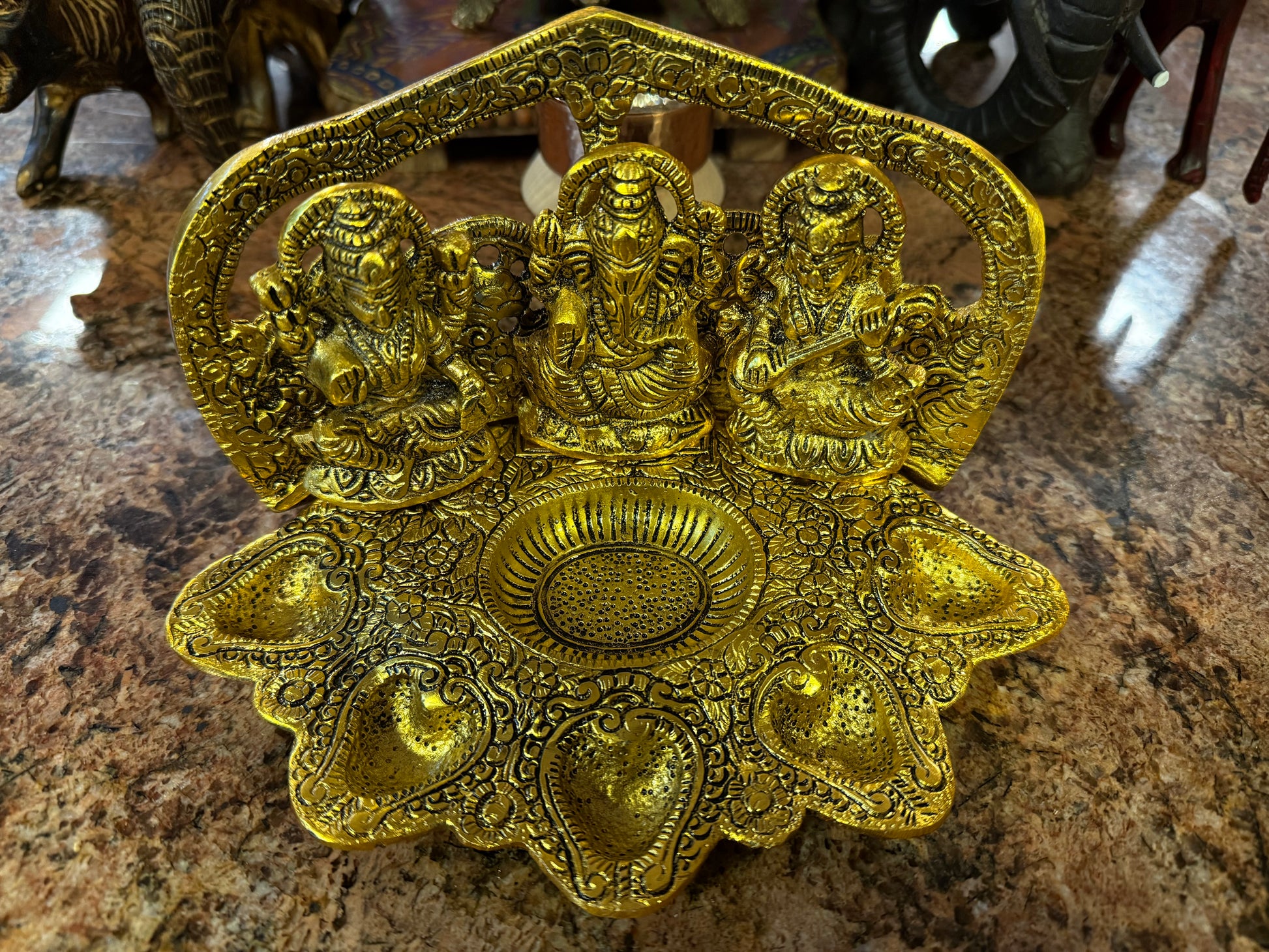 The Metal Lakshmi Saraswathi Ganesh Diya by Saanskrityam Handicraft Store is an ornate brass oil lamp featuring three seated deities. Its seven petal-like compartments and intricate carvings enhance its golden appearance, making it an ideal gift for special events.