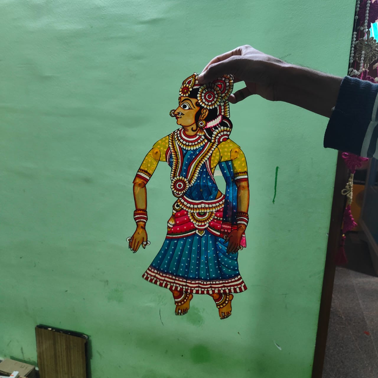 A hand displays a puppet from the Ramayana flat puppet set by Saanskrityam Handicraft Store, against a green wall. This elaborately decorated figure showcases vibrant colors—blue, red, and yellow—with intricate patterns and jewelry designs, potentially depicting the heroic Rama.