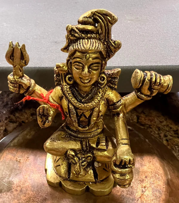 A 3 inch brass statue from Saanskrityam Handicraft Store depicts a Hindu deity, Shiva, with a trident and other objects, adorned with intricate jewelry. The figure displays a serene expression while sitting cross-legged on a small platform.