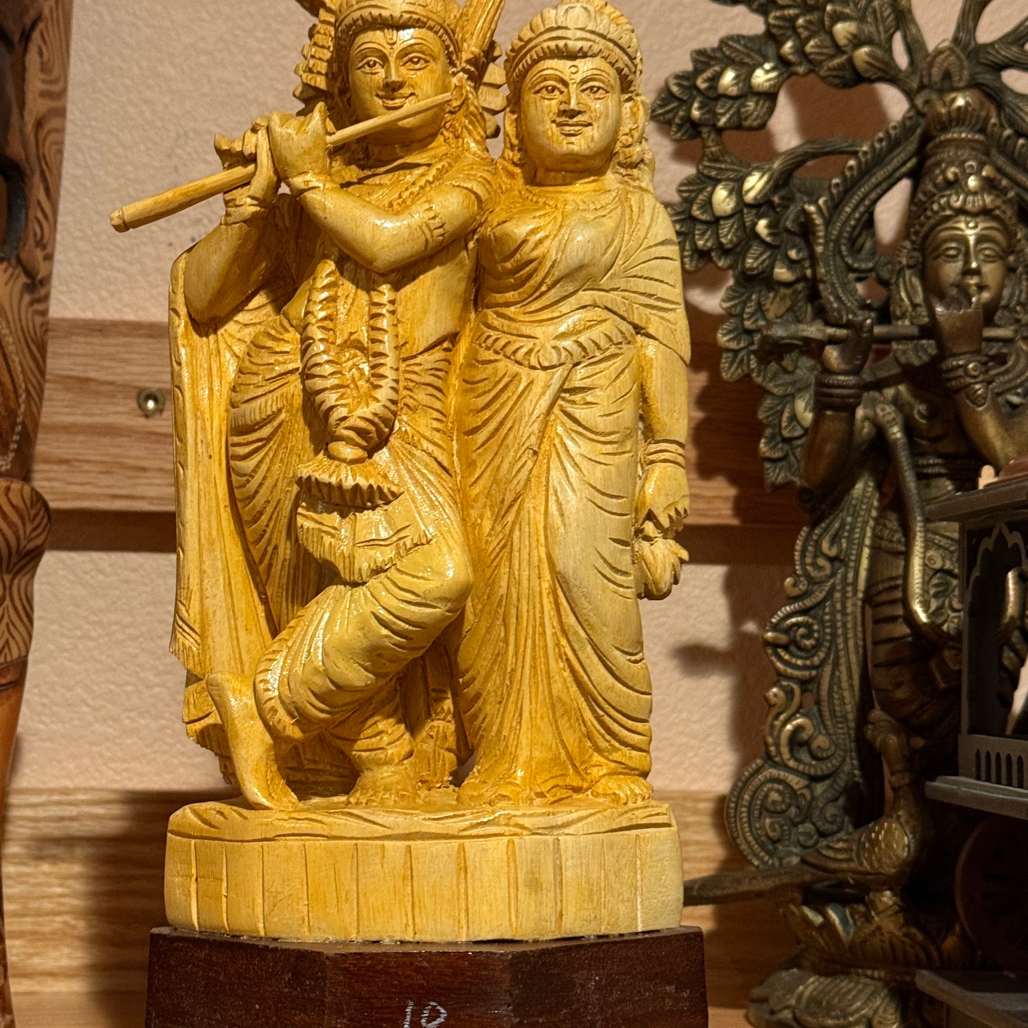 The Radhakrishna wood statue by Saanskrityam Handicraft Store features two handcrafted figures with one playing a flute, set on a wooden base. It stands out among other intricate sculptures on a shelf, offering a captivating collectible piece of art.