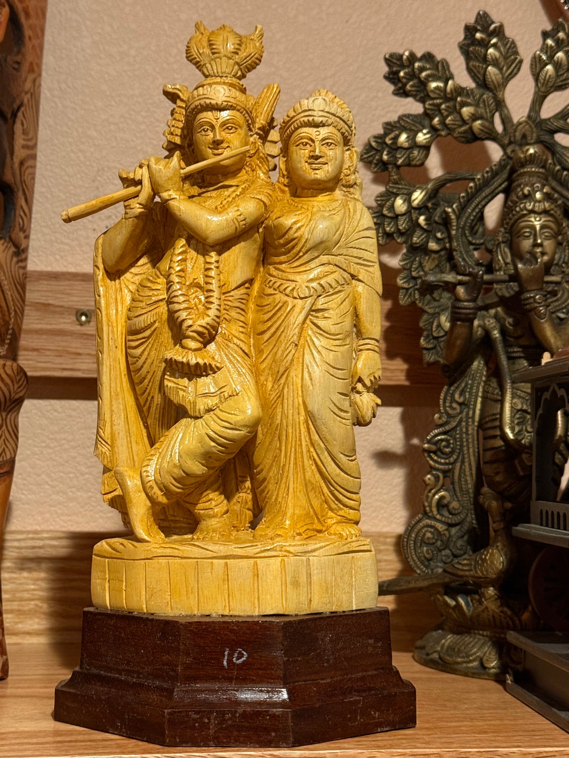 The Radhakrishna wood statue by Saanskrityam Handicraft Store features two handcrafted figures with one playing a flute, set on a wooden base. It stands out among other intricate sculptures on a shelf, offering a captivating collectible piece of art.