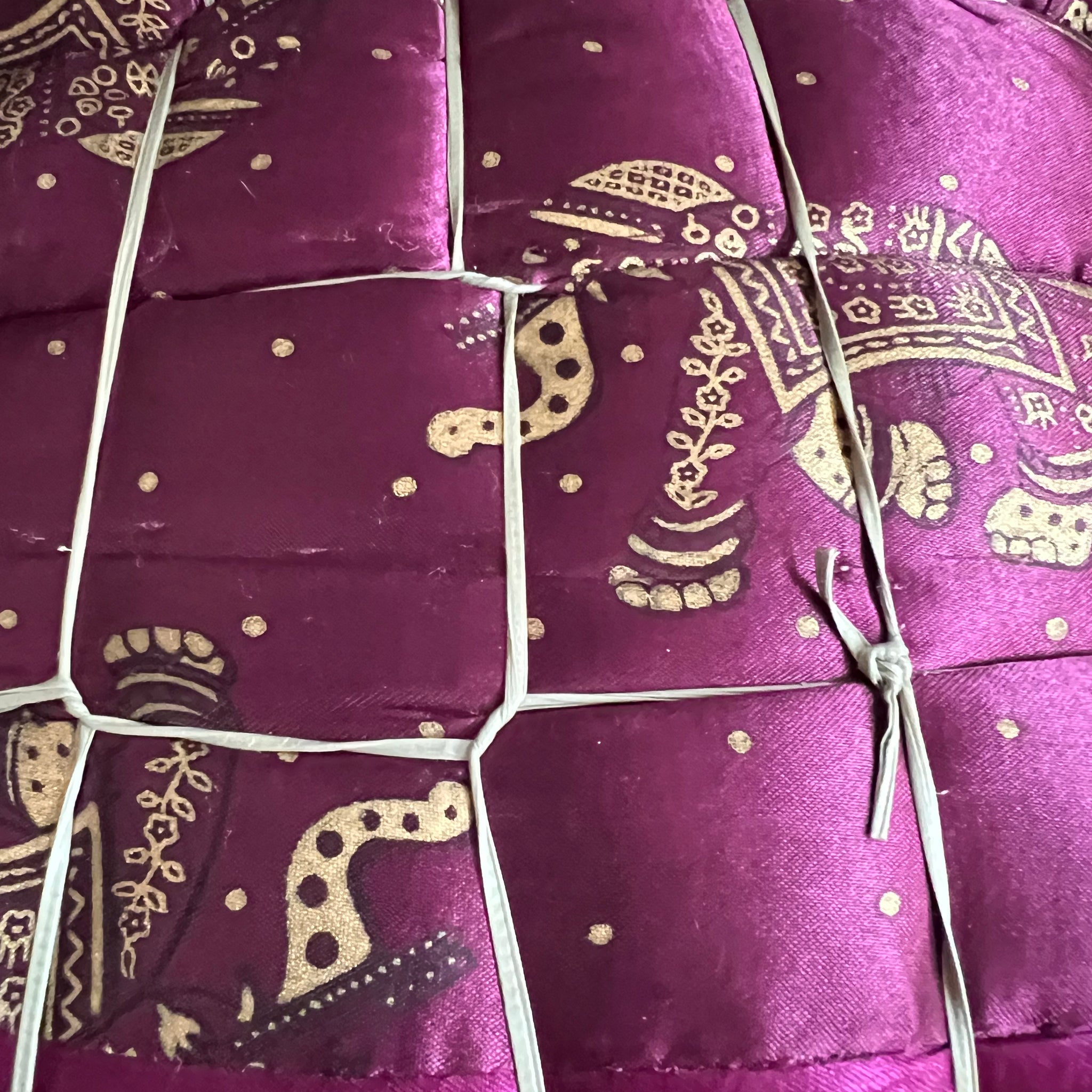 A close-up of the Jaipuri Razai, a cozy queen-size satin comforter by Saanskrityam Handicraft Store, featuring gold elephant designs on a purple background. This exquisite fabric, handmade in Rajasthan, is quilted and secured with white ties.