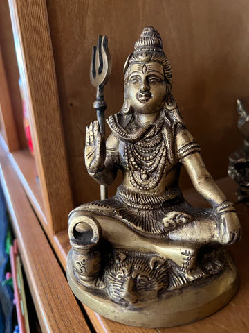 The 6-inch Shiva bronze statue by Saanskrityam Handicraft Store features a deity with a trident, seated in a meditative cross-legged position on a wooden shelf. The figure is intricately adorned with jewelry and exudes a calm expression, with a tiger depicted at the base of the statue.