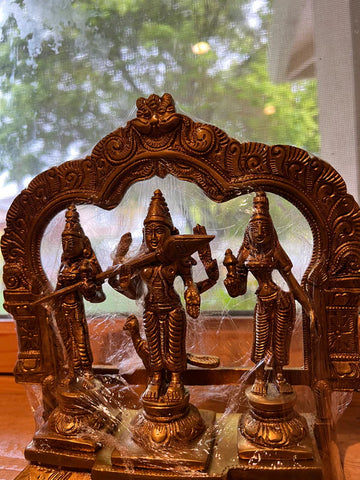 Intricate bronze statues from Saanskrityam Handicraft Store, featuring a 7-inch depiction of Kartikeya with consorts Valli and Devayani nestled in a stunning Prabhavali frame, are wrapped in clear plastic. They are presented against an outdoor backdrop softly blurred through a window.