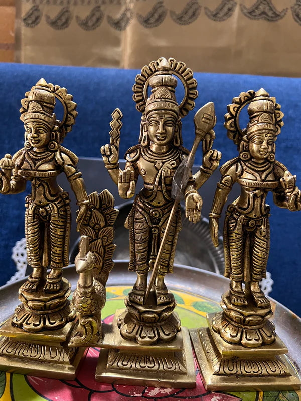 A set of three 8-inch bronze figurines depicting Kartikeya, Valli, and Devayani from the Saanskrityam Handicraft Store stands on a colorful tray. Each figure is intricately detailed with traditional attire and jewelry, with a small peacock statue placed in front of one deity. In the background, a blue couch can be seen.