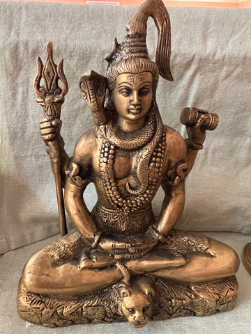 The 9 inch Shiva bronze in meditating pose from Saanskrityam Handicraft Store depicts a cross-legged Hindu deity holding a trident, featuring intricate details such as snakes and elaborate jewelry, and is displayed on a fabric surface.