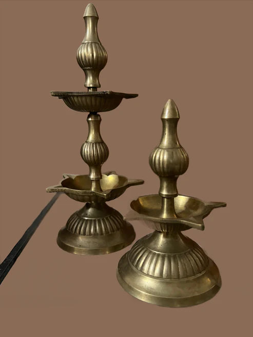 The Antique brass lamp set by Saanskrityam Handicraft Store, featuring intricately designed 7-inch and 4-inch brass oil lamps with a glossy finish, is placed on a surface. Each lamp showcases a tiered structure with ornamental detailing against a plain brown background.