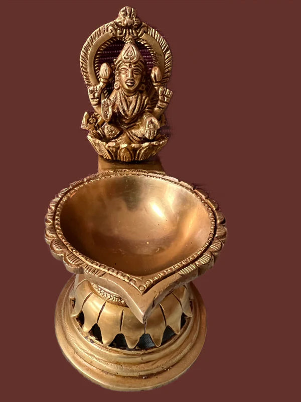 The Lakshmi lamp, crafted by Saanskrityam Handicraft Store, is a 7-inch-high brass oil lamp with an elaborate design featuring a seated deity figure on the back. It includes a wide, shallow bowl for holding oil and is set against a solid brown background.