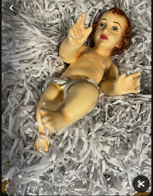 A unique Baby Jesus paper mache figurine, crafted by Saanskrityam Handicraft Store, features a baby in a cloth diaper resting on shredded white paper, with raised hands and a serene expression.