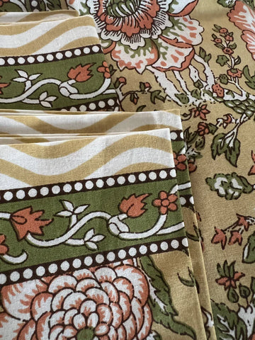 A detailed view of the Beige and Orange Print from Saanskrityam Handicraft Store reveals fabric adorned with complex floral and geometric patterns in shades of green, orange, and beige. It showcases large flowers, intertwining vines, and dotted borders, with several layers of folded fabric on display.