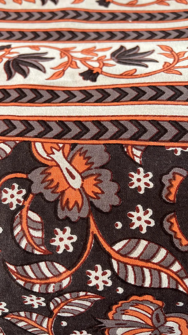 A close-up of the Brown and Orange Print from Saanskrityam Handicraft Store, showcasing floral and leaf motifs in vibrant orange and black, complemented by geometric stripes. The background features an intricate tapestry of beige, white, and black tones.