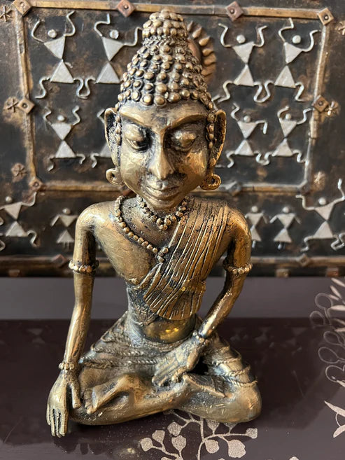 The Buddha bronze 9 inch statue from Saanskrityam Handicraft Store features a meditating figure with intricate details against a textured, decorative background. The statue is adorned with jewelry and a patterned robe, while the backdrop showcases geometric designs.