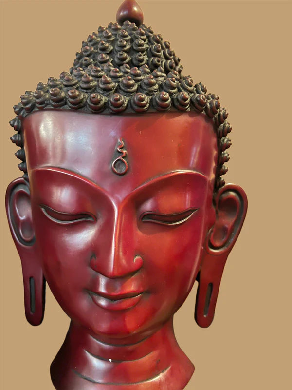 The image captures a close-up of the Buddha head mask red lapis from Saanskrityam Handicraft Store, featuring closed eyes, elongated earlobes, and intricately curled hair. The sculpture stands out against a plain, light brown background.