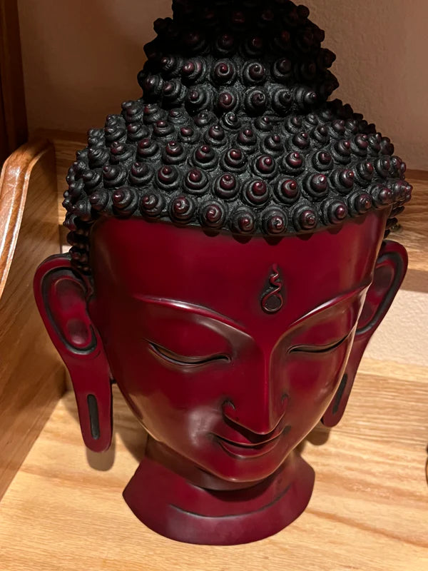 The Buddha head / mask red lapis from Saanskrityam Handicraft Store, featuring intricately detailed curly hair and a serene facial expression, rests on a wooden surface, contributing to a peaceful and contemplative ambiance.